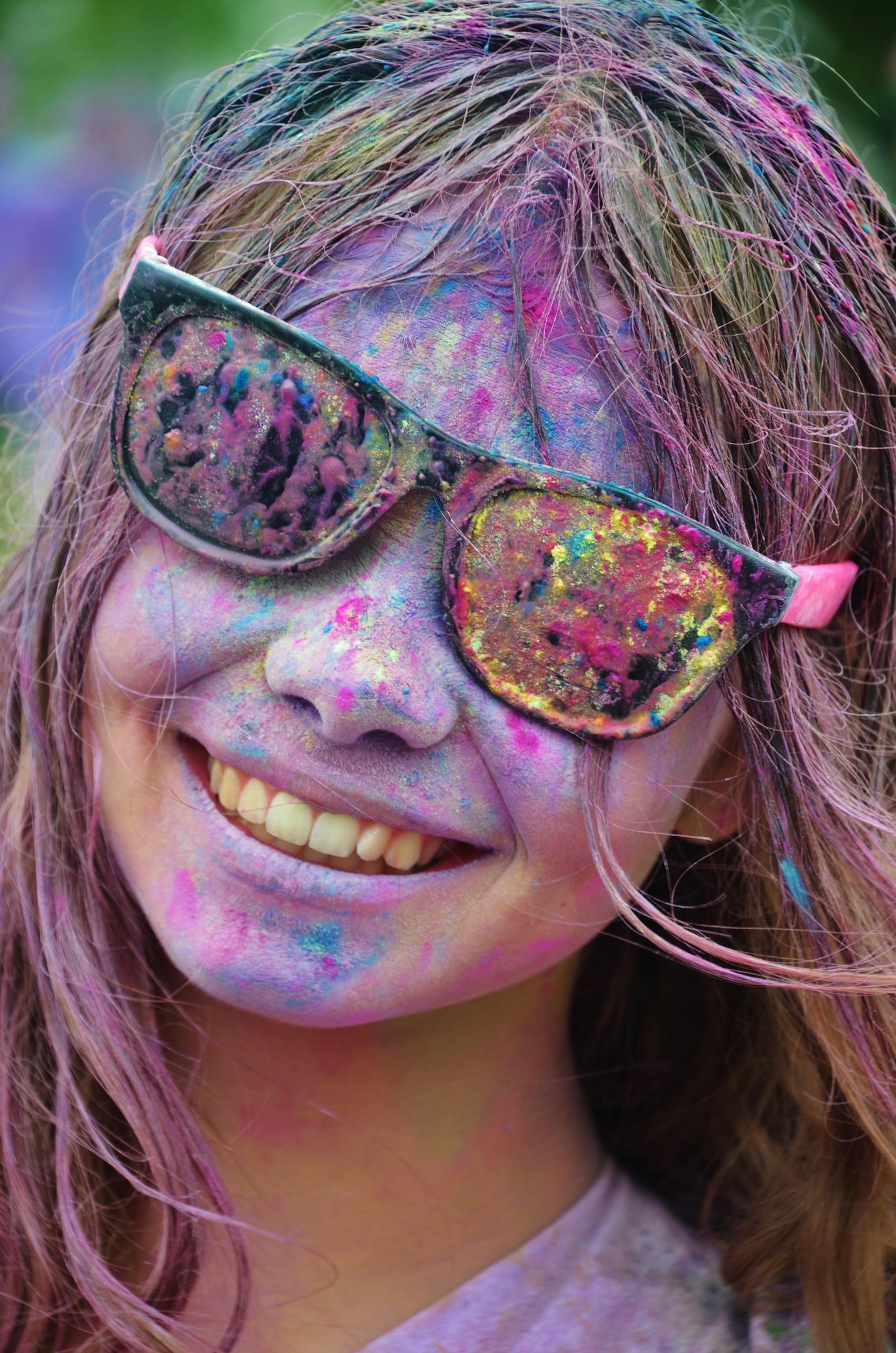 A smiling woman in sunglasses, covered with colorful powder at a festival celebrating joy and togetherness.